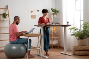 A woman working at a standing desk while a man works at a separate low desk using a stability ball, demonstrating active and ergonomic office setups.